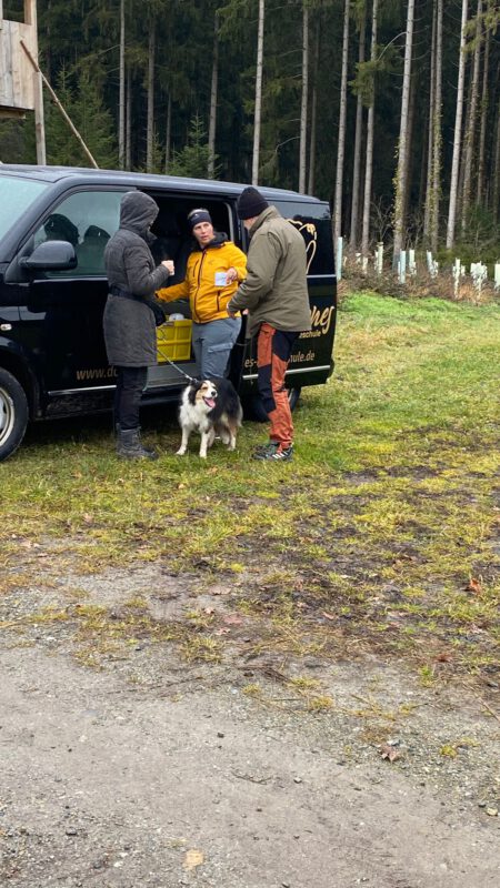 Trainerin der DogBones Hundeschule bespricht mit Teilnehmern den Ablauf des Socialwalks neben dem Trainingsfahrzeug im Wald, Hund steht entspannt an der Leine.